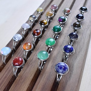 May include: A display of rings with various coloured gemstones, including opal, red jasper, green aventurine, and lapis lazuli. The rings are arranged on a wooden display stand.