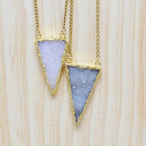 Puede incluir: Dos collares de cadena dorada con colgantes triangulares. Los colgantes están hechos de cuarzo transparente con un acabado brillante y brillante. Un colgante es rosa claro y el otro es gris claro.