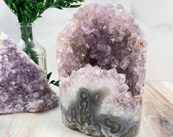 May include: A large amethyst geode with a natural opening, revealing a cluster of purple crystals. The geode has a smooth, gray exterior with a natural, rough texture.