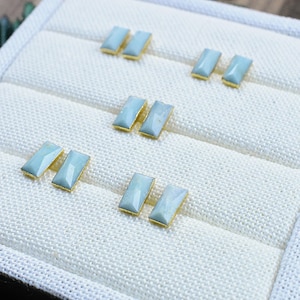 May include: A set of six light blue rectangular earrings with gold settings. The earrings are displayed on a white fabric background.