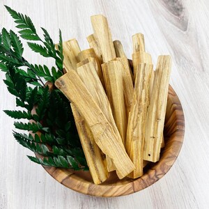 May include: A wooden bowl filled with light brown palo santo sticks and green fern leaves. The sticks have a natural wood grain pattern. The bowl is made of a light-colored wood. The arrangement is set against a light wood-grain background.