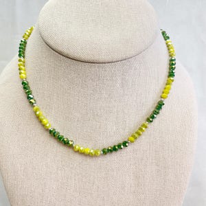 May include: A beaded necklace with a gradient of yellow and green faceted beads. The necklace is displayed on a neutral-colored bust form. The beads are strung on a silver-colored chain.