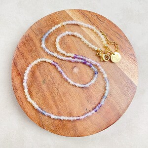 May include: Delicate beaded necklace with a gradient of colors, from white to purple. The necklace is displayed on a round wooden surface. The clasp and chain are gold-colored.