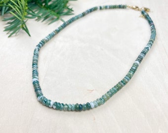 May include: A beaded necklace featuring small, round, moss agate stones in shades of green and white. The necklace has a gold-colored clasp and is displayed on a light-colored wooden surface. A sprig of green foliage is in the background.