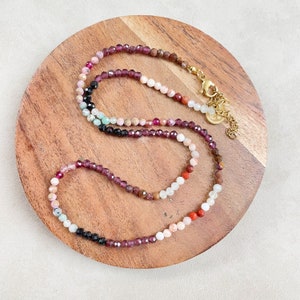 May include: A multi-colored gemstone necklace with a gold clasp. The necklace features a variety of stones, including pink, red, orange, brown, black, and white.