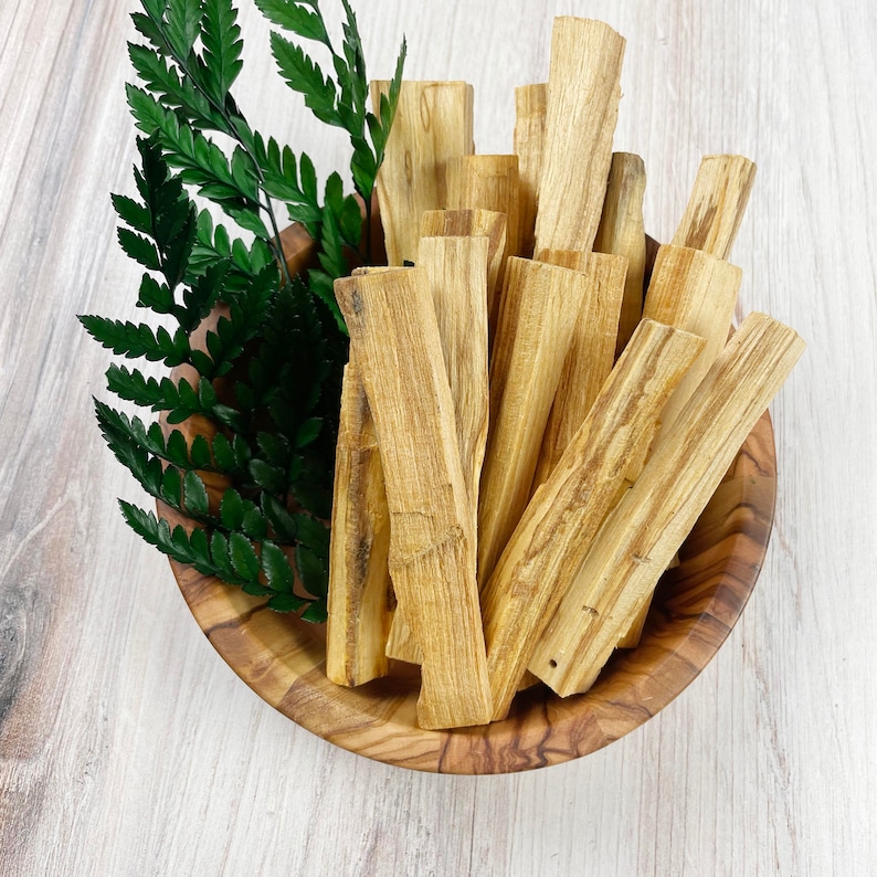 May include: A wooden bowl filled with light brown Palo Santo sticks and green fern leaves. The Palo Santo sticks are rectangular and have a natural wood grain. The bowl is made of wood and has a natural finish.