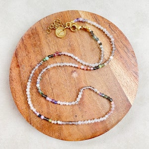 May include: A delicate necklace with a gold clasp and a variety of colorful gemstone beads. The beads are small and round, and they are strung on a thin chain. The necklace is displayed on a wooden surface.