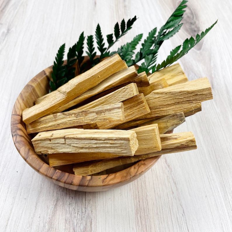 May include: A wooden bowl filled with light brown palo santo sticks and green fern leaves. The bowl has a natural wood grain pattern. The sticks are rectangular and have a textured surface. The arrangement is on a light wood surface.
