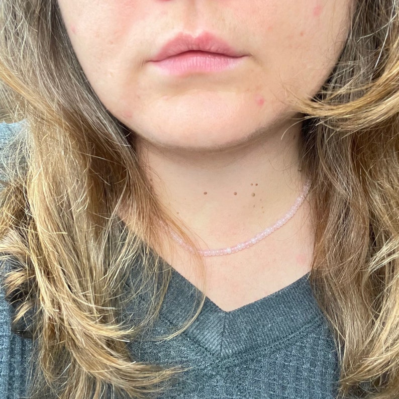 May include: A close-up of a person wearing a light gray v-neck shirt with a delicate pink beaded necklace.