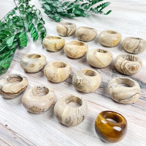 May include: A collection of 14 smooth, round, beige and brown banded rings made of natural stone. The rings are arranged on a light wood surface. One ring is a golden brown tiger's eye.