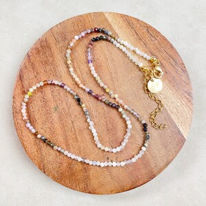 May include: A delicate multi-colored gemstone necklace with a gold clasp and a small round charm. The necklace is displayed on a wooden surface.