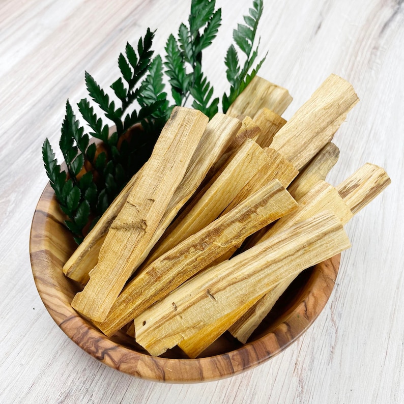 May include: A wooden bowl filled with light brown palo santo sticks. Green fern-like leaves are tucked around the sticks. The bowl has a natural wood grain pattern. The sticks have a light, natural color with visible wood grain.