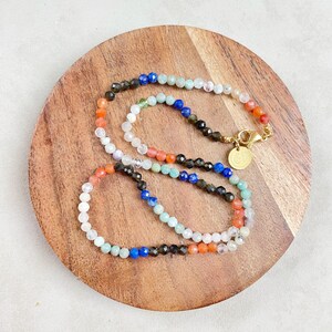 May include: A multi-strand necklace made of various gemstone beads. The necklace features beads in shades of blue, green, orange, and white. The beads are strung on a gold chain with a gold clasp.