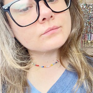 May include: A close-up of a person wearing a colorful beaded choker necklace. The necklace is made of small, multi-colored beads and is worn on a light blue shirt.