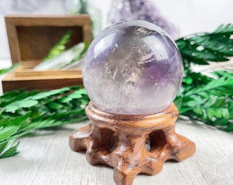 Op de afbeelding: Een paarse amethist kristallen bol op een houten standaard. De bol is helder en heeft een lichtpaarse tint. De standaard is gemaakt van donker hout en heeft een eenvoudig ontwerp.