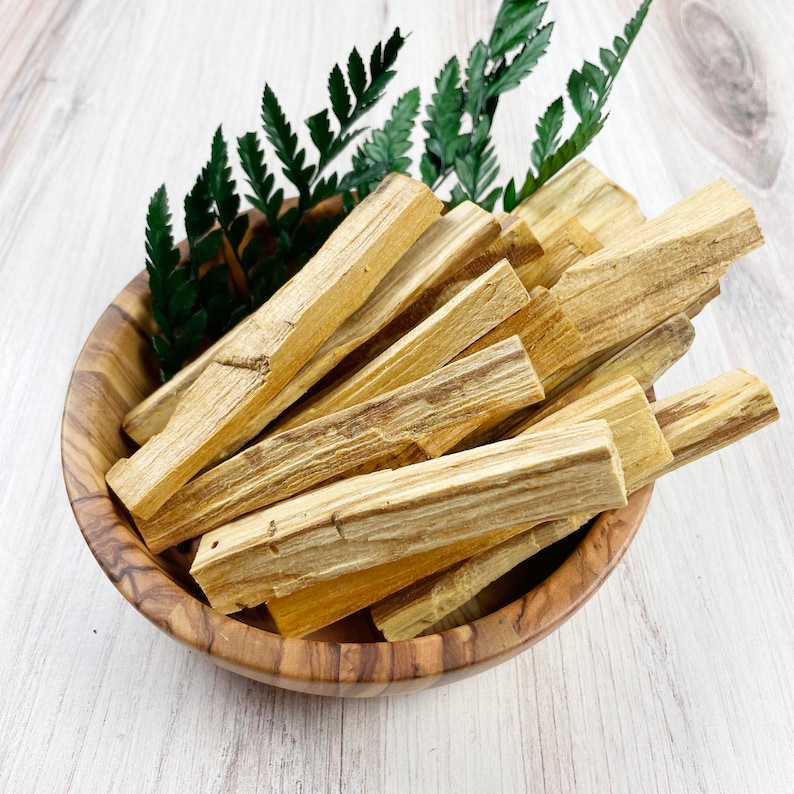 May include: A wooden bowl filled with Palo Santo sticks and green fern leaves. The Palo Santo sticks are light brown with darker brown streaks. The bowl is made of wood and has a natural wood grain pattern.
