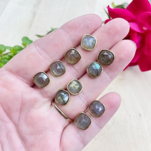 May include: Eight pairs of gold-toned earrings with square-shaped labradorite gemstones. The earrings are displayed on a hand.