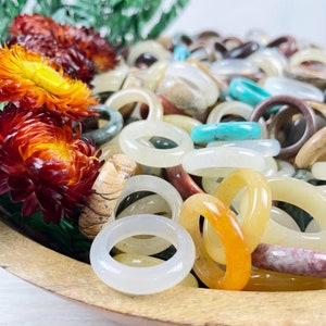 May include: A bowl filled with a variety of colorful stone rings. The rings are made of different types of stones, including white, brown, yellow, and turquoise. The rings are all different sizes and shapes.