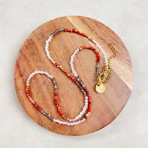 May include: A multi-colored beaded necklace with a gold clasp. The necklace is made of small, round beads in shades of red, pink, and gray. The beads are strung on a thin, clear cord. The necklace is displayed on a wooden surface.
