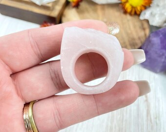 Pode incluir: Um anel de pedra natural rosa claro, talhado em bruto. O anel é feito de uma única pedra e tem uma forma simples e orgânica.