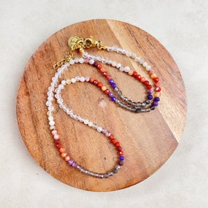 May include: A multi-strand necklace with a gradient of gemstone beads. The necklace features clear, pink, red, orange, purple, and gray beads.