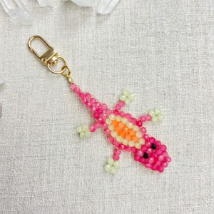 May include: A beaded lizard keychain in shades of pink, orange, and green. The lizard has a gold-colored clasp and is made of small, colorful beads. The keychain is a decorative accessory.