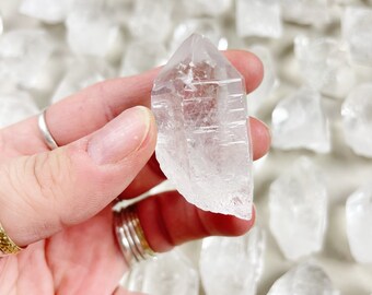 Peut inclure: Une collection de pointes de cristal de quartz clair, avec une pointe tenue dans une main. Les pointes sont de tailles et de formes variées, certaines ayant une pointe plus pointue que d'autres.