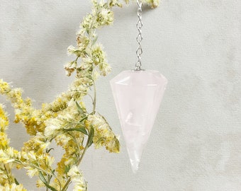 Puede incluir: Un péndulo de cristal de cuarzo rosa colgado de una cadena plateada. El péndulo tiene forma de cono puntiagudo con un diseño facetado. El cristal es translúcido y tiene un tono rosa claro.