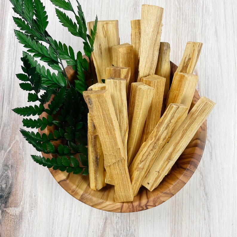May include: A wooden bowl filled with Palo Santo sticks and green fern leaves. The Palo Santo sticks are light brown and have a natural, textured appearance. The bowl is made of wood and has a round shape. The fern leaves are a vibrant green.