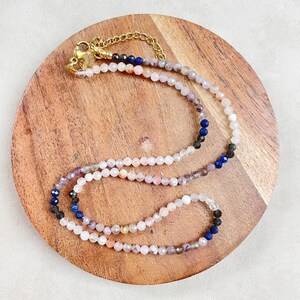 May include: A multi-colored beaded necklace with a gold clasp. The beads are pink, white, blue, and black. The necklace is on a wooden surface.