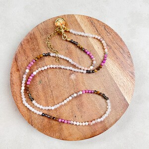 May include: A delicate layered necklace with pink, brown, and white beads. The necklace has a gold clasp and is displayed on a wooden surface.