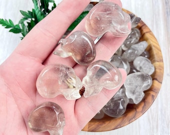 May include: A collection of clear quartz crystal skulls. The skulls are smooth and polished, with a translucent appearance. They are arranged in a bowl, with some of the skulls being held in a hand.