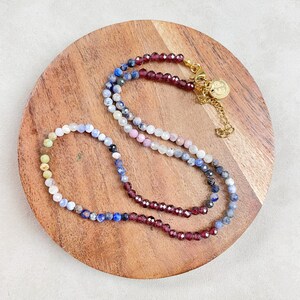 May include: A multi-colored gemstone beaded necklace with a gold clasp. The necklace features a gradient of colors, starting with yellow and transitioning to blue and then red.