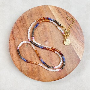 May include: A multi-strand necklace with small, colorful gemstone beads. The necklace is made of brown, blue, white, and pink beads. The necklace has a gold clasp and a small gold charm.