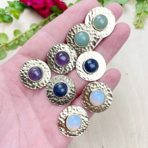 Pode incluir: Sete pingentes redondos de metal dourado com pedras preciosas de diferentes cores no centro. As pedras preciosas são ametista, lápis-lazúli, opala e aventurina verde.