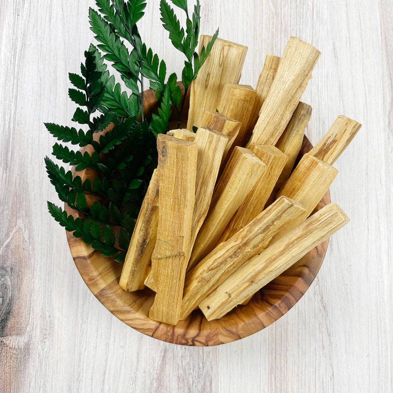 May include: A wooden bowl filled with Palo Santo sticks and green fern leaves. The Palo Santo sticks are light brown with visible wood grain. The bowl is made of olive wood and sits on a white wooden surface.