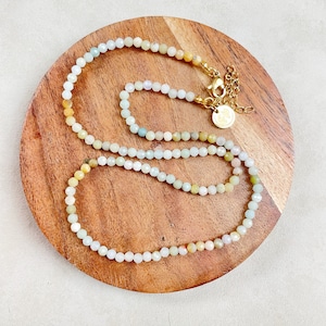 May include: A delicate necklace made of small, faceted beads in shades of yellow, green, and blue. The necklace is layered on a wooden surface and features a gold clasp.