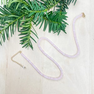 May include: A delicate pink rose quartz necklace with a gold clasp and a small extender chain. The necklace is made of small, round beads and is displayed on a light wood surface.