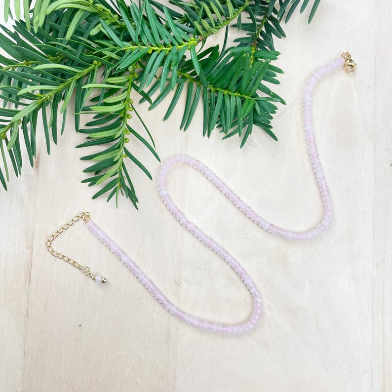 May include: A delicate pink rose quartz necklace with a gold clasp and a small extender chain. The necklace is made of small, round beads and is displayed on a light wood surface.