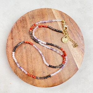 May include: A multi-colored beaded necklace with a gold clasp and a small gold tag with the letters "EP" on it. The necklace is made of small, round beads in shades of red, orange, purple, and gray. The necklace is lying on a wooden surface.