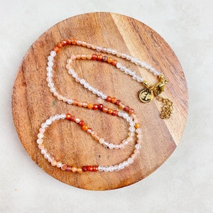 May include: A multi-strand necklace made of small, round beads in shades of white, orange, and red. The necklace has a gold clasp and is displayed on a wooden surface.