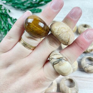May include: A hand wearing a gold ring is holding three rings made of natural stone. The rings are made of tiger's eye and banded agate. The rings are a light brown color with brown and black stripes.