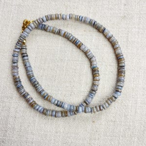 Può includere: Una collana composta da piccole perline rotonde, azzurre e beige. Le perline sono infilate insieme, formando una delicata catena. La chiusura è color oro. La collana è esposta su una superficie di colore neutro.