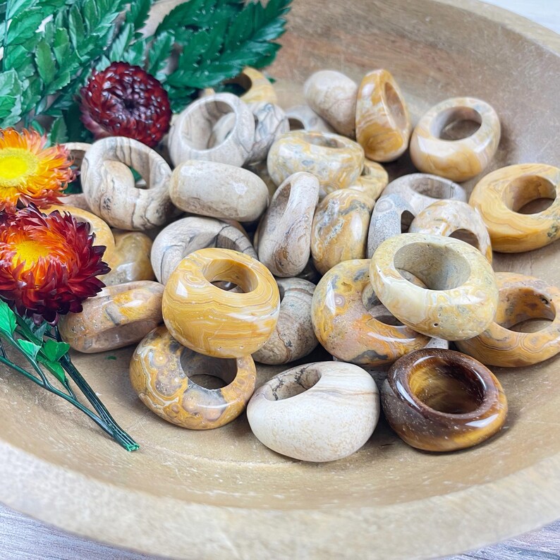 May include: A bowl filled with a variety of beige and brown agate rings. The rings are smooth and polished, with a natural, earthy look.