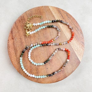 May include: A delicate beaded necklace with a variety of colorful gemstones. The necklace features a gold-colored clasp and extender chain. The beads transition from light blue to red and brown tones.