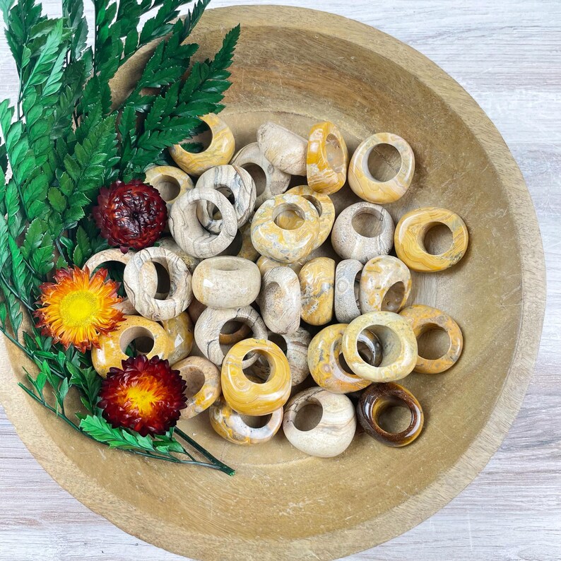 May include: A bowl filled with a variety of yellow and brown agate stone rings. The rings are smooth and polished, and they have a natural, organic look.