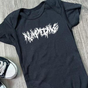 Puede incluir: Body de bebé negro con la palabra "NAPALM" en letras blancas, estilizadas y dentadas. El body está junto a un par de zapatillas de bebé en blanco y negro.