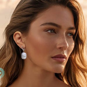 Puede incluir: Una mujer lleva un pendiente de plata con una perla blanca cuadrada y grande. El pendiente cuelga de un aro de plata. La mujer tiene el pelo largo y ondulado de color marrón y mira hacia un lado. El fondo es una vista borrosa del mar y el cielo.