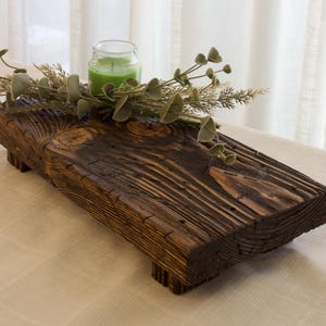 May include: A rustic, rectangular wooden centerpiece with a dark brown finish. It features a small green candle in a glass jar, and is decorated with faux greenery. The centerpiece is placed on a light-colored surface.