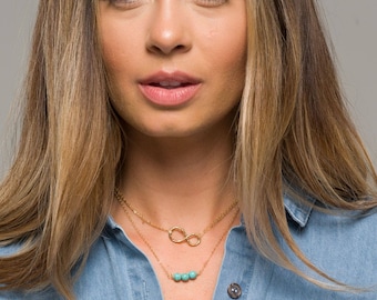 Puede incluir: Una mujer lleva una camisa vaquera azul claro con dos collares de oro. Uno de los collares tiene un símbolo de infinito y el otro una fila de cuentas turquesa.